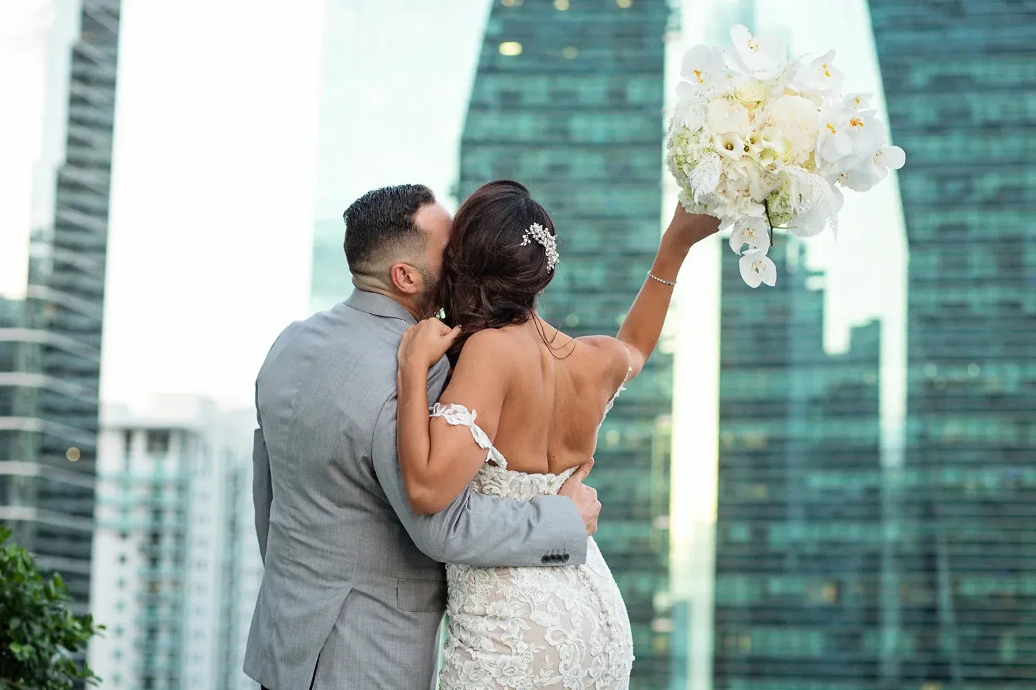 A bride in a stunning lace gown holds a beautiful bouquet while embracing her groom against a city skyline.