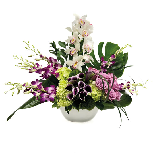 Elegant floral arrangement featuring white orchids, purple calla lilies, roses, and vibrant greenery in a modern vase.