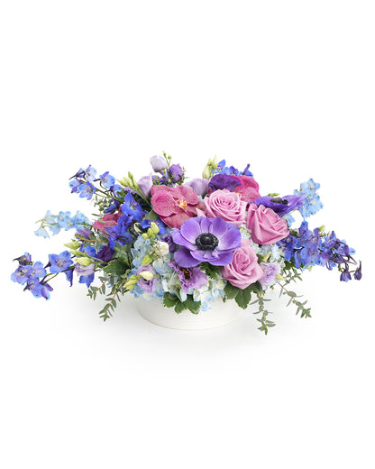 A vibrant floral arrangement featuring purple roses, orchids, and delphiniums in a stylish white vase.