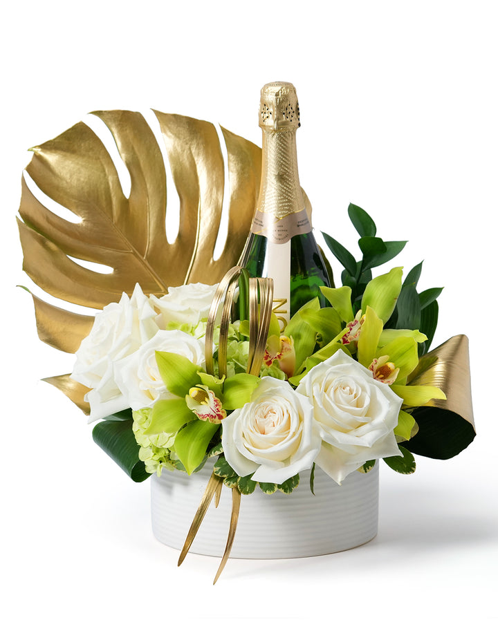 Elegant floral arrangement featuring white roses, green orchids, and a sparkling champagne bottle, accented with gold leaves.