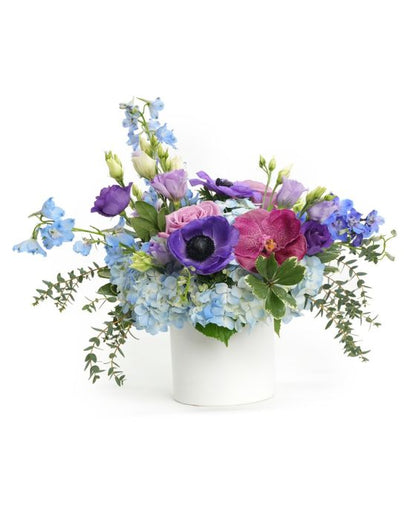 A vibrant floral arrangement featuring blue hydrangeas, purple orchids, and leafy accents in a white vase.